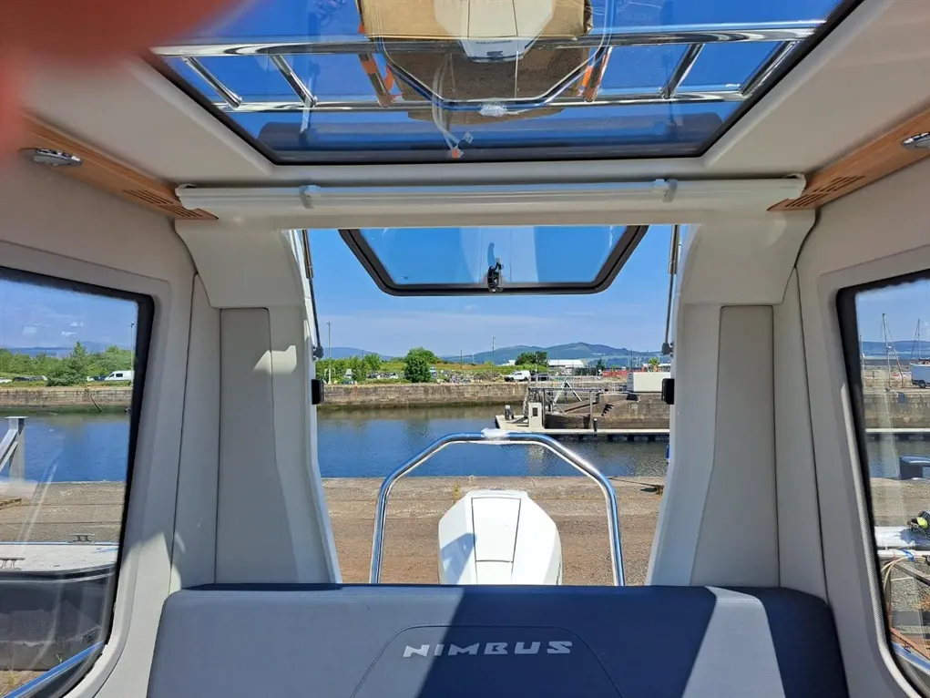 View from inside a boat cabin, featuring a clear sky, a canal with moored boats, and the word "NIMBUS" on the seat.